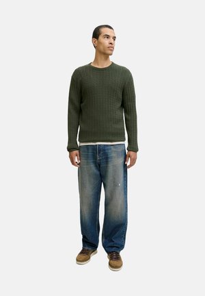 Man in olijfgroene kabeltrui, met losse blauwe jeans en bruine schoenen, staande tegen een effen lichte achtergrond, kijkend naar rechts.