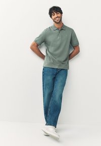 Grön pikétröja med krage, kombinerad med ljusblå jeans och vita sneakers. Modellen poserar mot en enfärgad bakgrund med en avslappnad hållning.