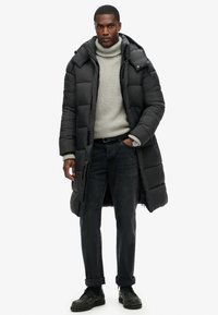 Abrigo de plumas negro con capucha, con textura acolchada y un diseño largo. Se lleva sobre un cuello de tortuga gris claro y jeans oscuros.