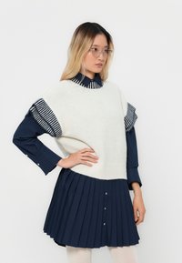 Weißer Strickpullover mit kurzen Ärmeln in Navy-Streifen, kombiniert mit einem plissierten, dunkelblauen Hemd. Mit Knopfdetails und geschichtetem Design.