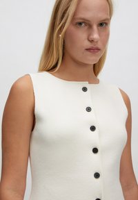 Weißes ärmelloses Kleid mit strukturiertem Oberflächenmaterial und Rundhalsausschnitt, das fünf schwarze Knöpfe auf der Vorderseite hat.