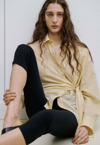 Mladi človek z dolgimi kodrastimi lasmi sedi ob steni, oblečen v rumeno srajco z gumbi, zavezano v pasu, in črne pajkice.