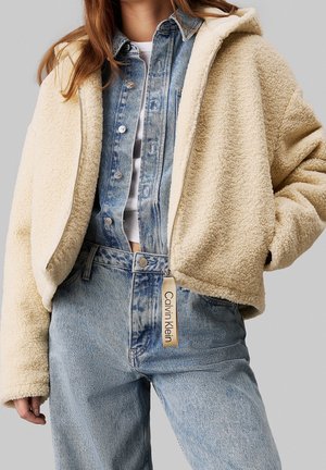 Femme portant une veste sherpa beige zippée par-dessus une veste en jean bleue et un jean avec la tirette de fermeture éclair Calvin Klein visible.