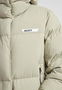 Lys beige pufferjakke med høj krave, lynlåslomme og hvidt logo-patch med teksten "JACK1T" på forsiden. Glat tekstur.