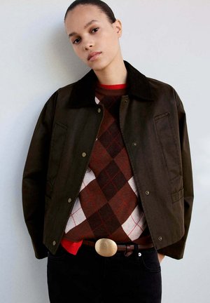 Joven con una chaqueta marrón oscuro sobre un suéter argyle rojo y marrón con una hebilla de cinturón grande y redonda, posando frente a un fondo liso.