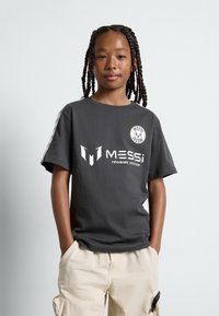 Camiseta de manga corta gris hecha de algodón, con gráficos blancos que incluyen "MESSi" y un logo circular en el pecho, combinada con pantalones cortos tipo cargo beige.