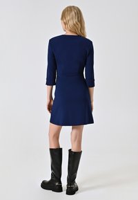 Marineblaues Kleid mit Dreiviertelärmeln und tailliertem Schnitt, das am Saum ausgestellt ist. Kombiniert mit knielangen schwarzen Lederstiefeln.
