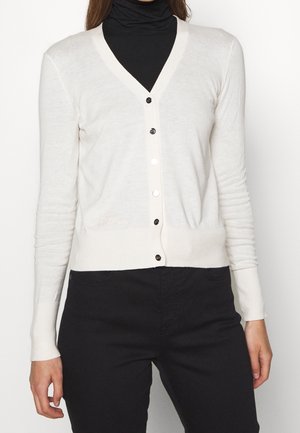 Kobieta ubrana w dopasowany biały sweter na guziki, nałożony na czarny golf i czarne spodnie, pokazana od ramion do bioder.