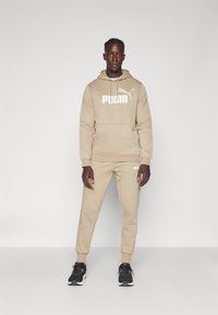 Beige Trainingsanzug mit Kapuzenpullover und großem weißen Puma-Logo sowie passenden Jogginghosen. Das Material wirkt weich und strukturiert.