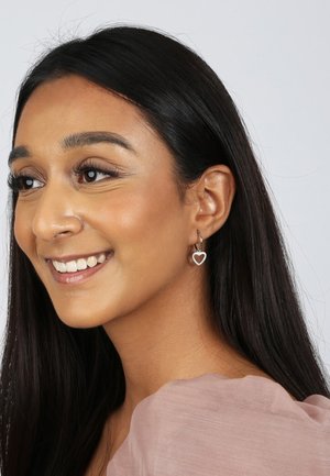 Boucles d'oreilles en argent en forme de créole avec un charm en forme de cœur, orné de petites pierres claires, suspendu au lobe de l'oreille.