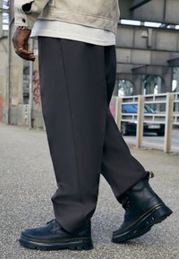 Botas de cuero negras con suelas texturizadas y costuras amarillas, combinadas con pantalones sueltos de color gris oscuro. Entorno urbano con fondo de concreto.