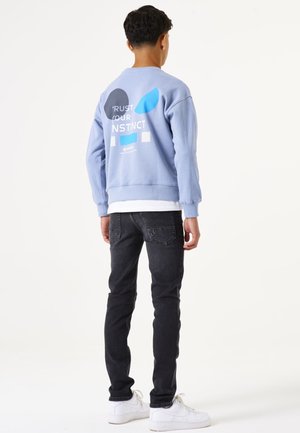 Lichtblauwe sweatshirt met grafische tekst en vormen op de achterkant, gecombineerd met donkere skinny jeans en witte sneakers. Minimalistisch ontwerp.