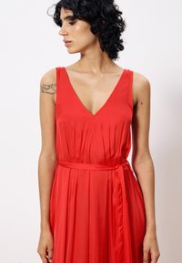 Red sleeveless dress with a V-neckline, pleated design, and a tie belt at the waist. Smooth fabric with a flowing silhouette.