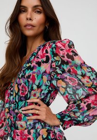 Frau mit langen braunen Haaren trägt eine bunte Bluse mit Puffärmeln im Blumenmuster und einen goldenen Ring, posiert vor einem einfarbigen, hellen Hintergrund.