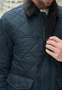 Gestepptes marineblaues Jackett mit einem Cordkragen, Reißverschluss vorne und Druckknopfverschluss, aus einem strukturierten Stoff und mit zwei Fronttaschen.