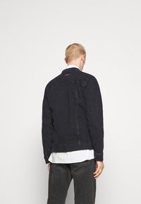 Giacca di jeans scura con una vestibilità diritto, dotata di colletto, maniche lunghe e dettagli di cucitura visibili. Indossata sopra una camicia bianca.