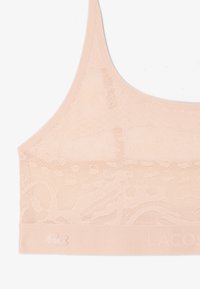 Bralette en dentelle rose clair avec des motifs floraux complexes, bretelles fines et une large bande ornée d'un petit logo crocodile brodé.