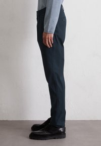 Donkergroene, rechte broek van een katoenmix, met een zijzak en een elastische tailleband. Gepaard met zwarte veterschoenen.