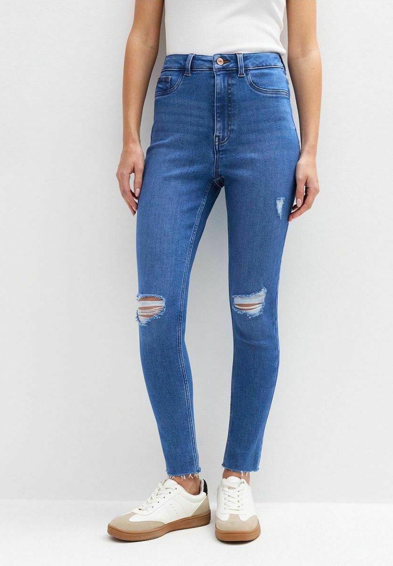 Jeans de tiro alto en azul con un corte ajustado, que presentan desgarros en las rodillas y dobladillos deshilachados, combinados con zapatillas blancas y beige.