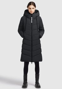 Lang sort dunjakke med hætte, prydet med chevron-syninger, lynlås lukning og snorede detaljer. Bæres med sorte leggings og støvler.