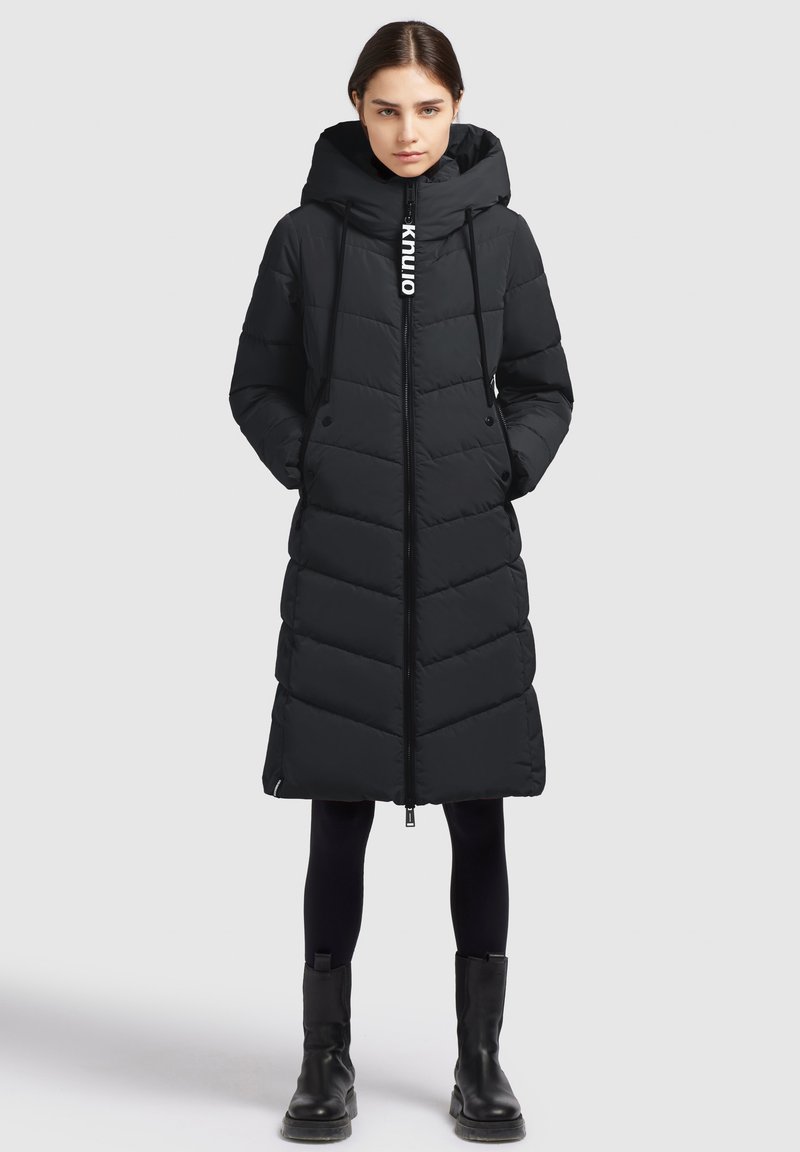 Lang sort dunjakke med hætte, prydet med chevron-syninger, lynlås lukning og snorede detaljer. Bæres med sorte leggings og støvler.