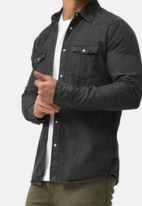 Schwarze Jeanshemd mit zwei Brusttaschen, Button-Down-Kragen und Knöpfen auf der Vorderseite, über einem weißen T-Shirt getragen, kombiniert mit olivgrünen Hosen.