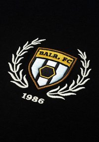 Wyhaftowane logo BALR. FC na czarnym materiale, w kształcie tarczy z złotymi, białymi i niebieskimi akcentami, z rokiem "1986" poniżej.