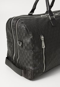 Bolsa de viaje negra con textura que presenta dobles cremalleras plateadas, asas de cuero y una base negra sólida con acentos decorativos en la superficie.