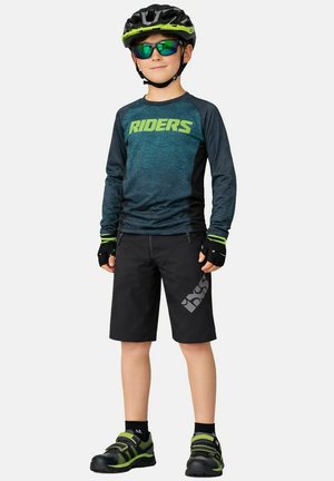 Junge trägt einen grün-schwarzen Fahrradhelm, Sonnenbrille, ein langärmeliges Radtrikot mit der Aufschrift "RIDERS", schwarze Shorts, Handschuhe und Schuhe mit grünen Akzenten.