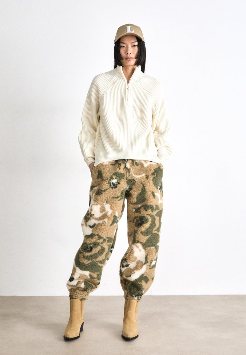 Kremowy sweter w prążki z pół zamkiem, w połączeniu z pluszowymi joggerami w beżowo-zielony kamuflaż i tanimi botkami za kostkę. Czapka z wyraźnym logo 'L'.