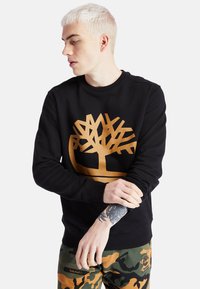 Felpa nera con un grande logo di un albero dorato, colletto rotondo, maniche lunghe e polsini a coste. Il modello la abbina a pantaloni camouflage.