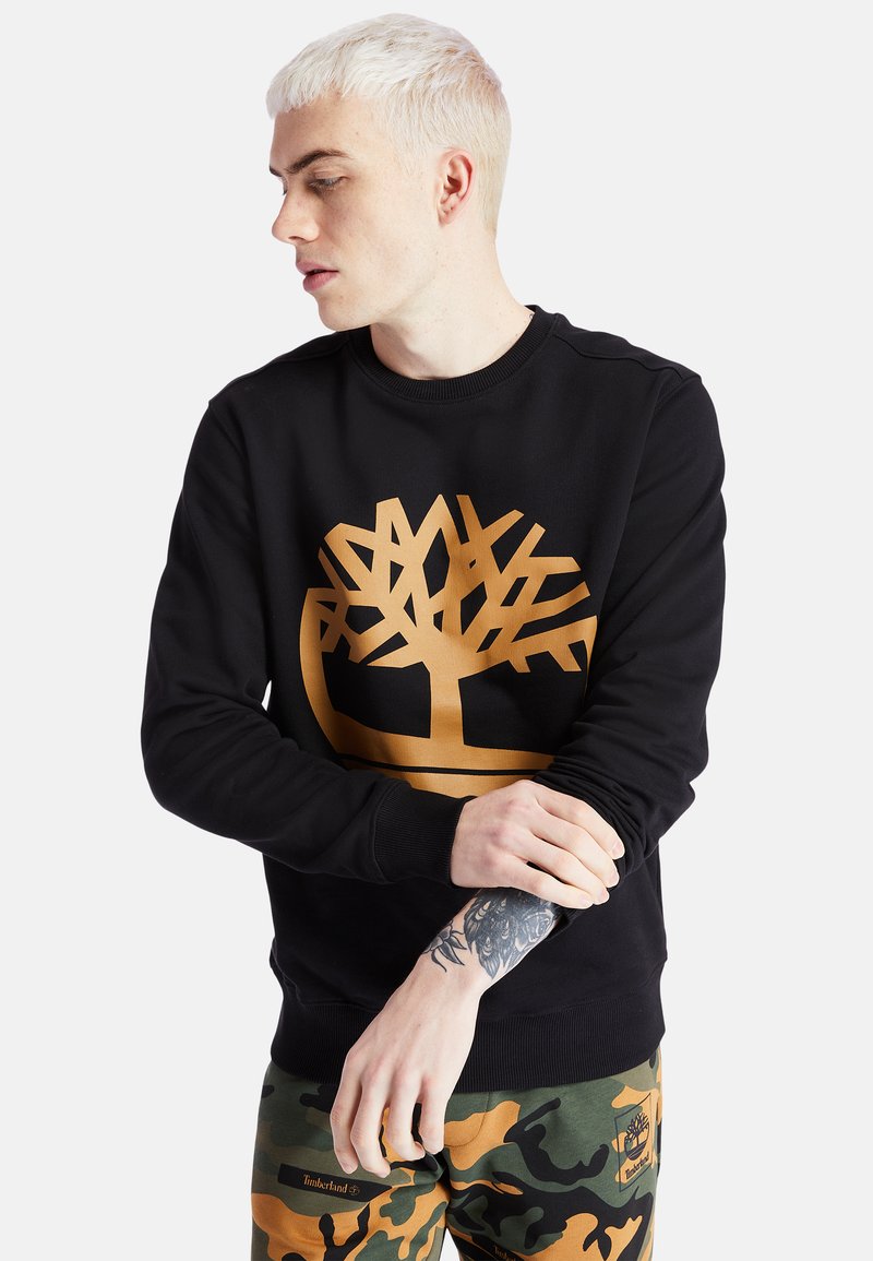 Felpa nera con un grande logo di un albero dorato, colletto rotondo, maniche lunghe e polsini a coste. Il modello la abbina a pantaloni camouflage.