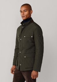 Maslinasto zelena jakna s preklopom, visokim ovratnikom, dva prednja džepa i srebrnim hardverskim detaljima. Teksturalna tkanina s krojem prilagođenim tijelu.