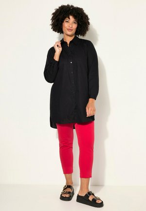 Schwarzes Langarmshirt mit Knopfverschluss, kombiniert mit knöchellangen leuchtend roten Hosen und schwarzen Sandalen mit goldenen Akzenten.