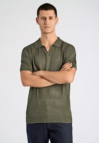 Kortärmad polo i olivgrön, tillverkad av textil med struktur. Har en V-ringad krage och ribbad nederkant, kombinerad med mörka byxor.