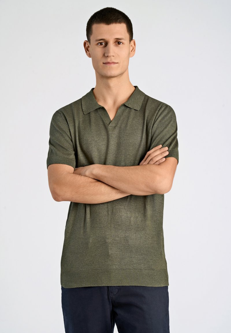 Kortärmad polo i olivgrön, tillverkad av textil med struktur. Har en V-ringad krage och ribbad nederkant, kombinerad med mörka byxor.