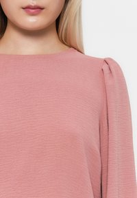 Rosa teksturert topp med rund hals og myke plisserte detaljer på skuldrene, laget av et lett og flytende stoff.