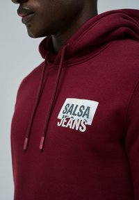 Zbliżenie na osobę w bordowej bluzie z kapturem z nadrukiem "SALSA JEANS" na piersi.