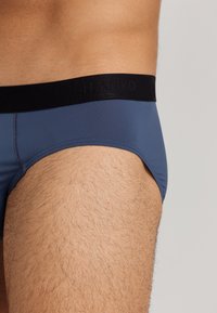 Boxer slip da uomo blu aderente con elastico nero in vita recante il nome del marchio "HANRO." Tessuto morbido e texture liscia con dettagli di cucitura minimi.