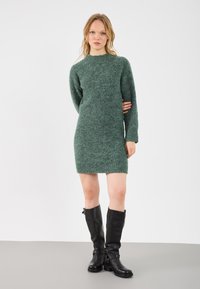 Grønn, lodden strikket kjole med lange ermer og rund hals, med hemline over kneet. Kombinert med høye svarte lærstøvler med spenner.