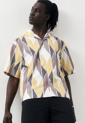 Mężczyzna o ciemnej karnacji i dredach, ubrany w krótką koszulę zapinaną na guziki z żółtymi, brązowymi i szarymi geometrycznymi wzorami oraz czarne spodnie.