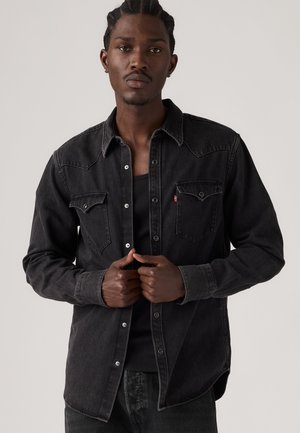 Mann mit geflochtenem Haar, der ein schwarzes Denim-Button-up-Hemd und ein schwarzes Tanktop trägt und das Hemd vorne zumacht.