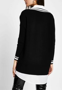 Zwarte geribbelde trui met een wit overhemd met kraag eronder, voorzien van gestreepte accenten op de manchetten. Eenvoudige, ontspannen pasvorm.