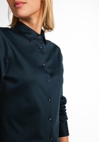 Camicia blu navy con collo a camicia, maniche lunghe e bottoni a contrasto, realizzata in un tessuto liscio con una leggera lucentezza.