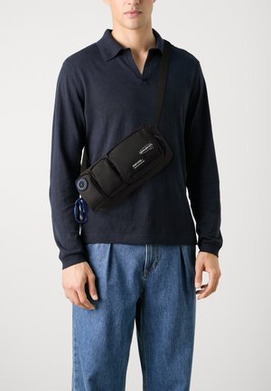 Svart axelväska med tre zippered fack, tillverkad av nylon, med en cirkulär detalj i metall och en nyckelringsklipp, bärs över kroppen.