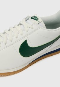 Tênis de couro branco com um logótipo Swoosh de couro verde, atacadores brancos e uma sola de borracha com textura em zigue-zague. Acento azul no calcanhar.