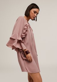 Rochie roz cu volane, cu mâneci lungi, realizată dintr-un material ușor. Designul include un guler legat cu un cordon și o croială lejeră, evidențiind o textură subtilă.