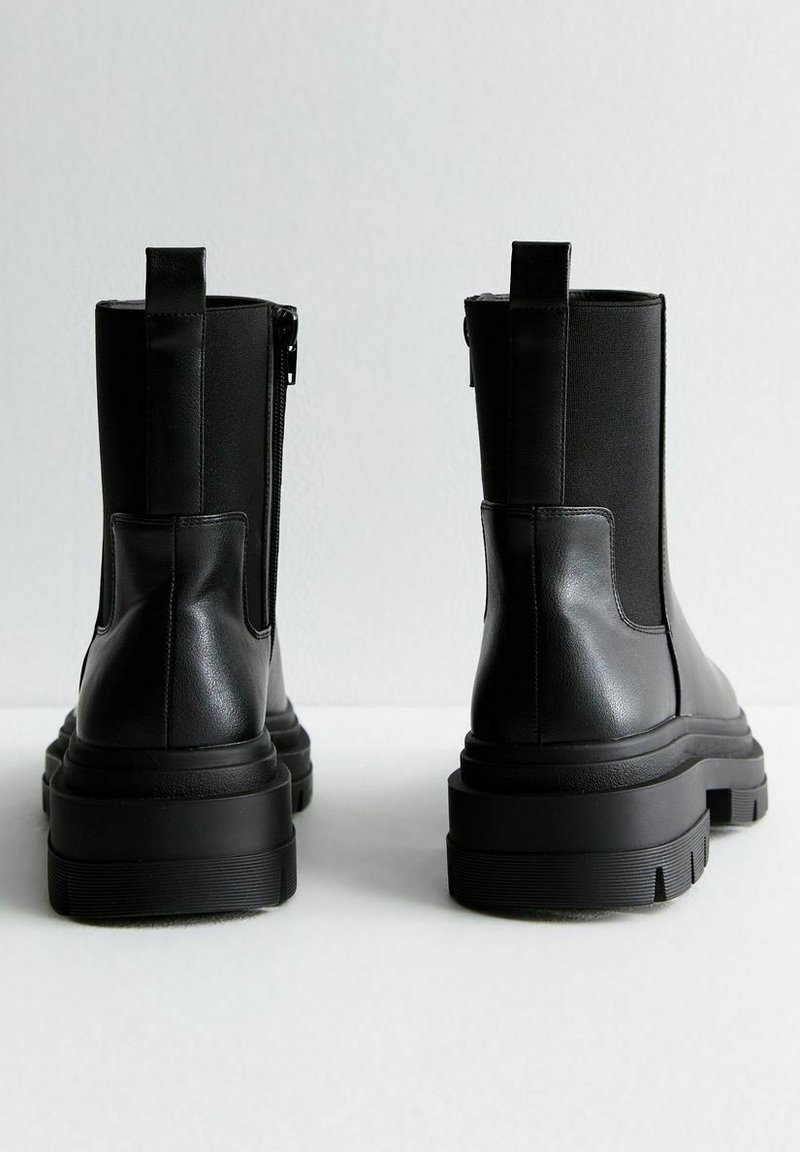 Une paire de bottines en cuir noir avec des panneaux latéraux en élastique et des semelles en caoutchouc épaisses, présentées de dos sur une surface blanche.