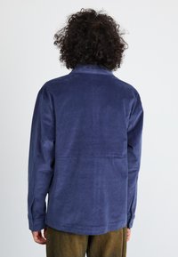 Navy blue corduroy shirt with a collar and buttoned cuffs, featuring a back yoke seam and a relaxed fit. Textured fabric visible.