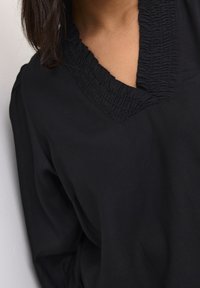 Haut noir avec un col smocké, un design en V, des manches longues et un tissu doux et texturé.